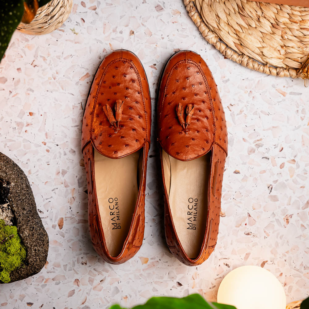 A pair of luxury men's footwear—Aubiere Cognac loafers by Marco Di Milano—boasts rich brown ostrich leather, with one shoe upright and the other on its side to reveal a sole stamped "Marco Di Milano.