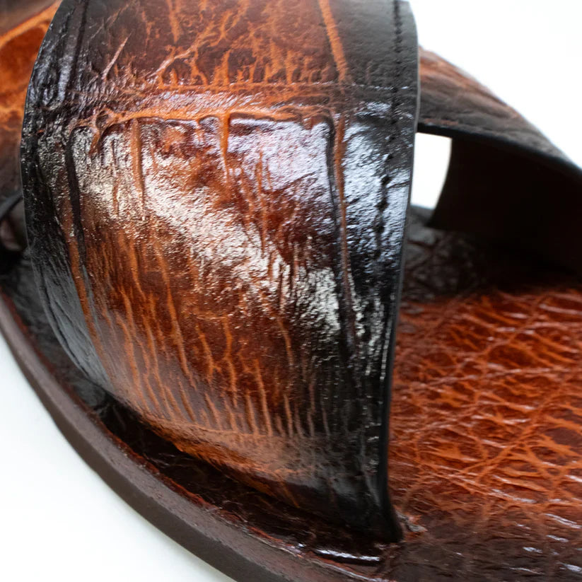 The Elios Honey by Marco Di Milano is a luxury brown leather sandal with a crocodile-textured finish and two crisscross straps, hand painted for elegance and showcased on a white background.