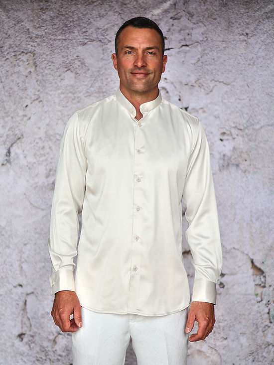 A man stands against a textured white wall, wearing the Manzini MANZINI INR-102 Banded Collar Solid Color Satin Cream Shirt and white pants.