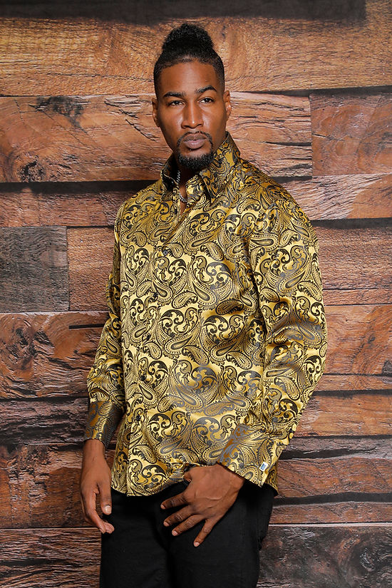 A man wearing the Manzini MANZINI MZT-100 Paisley Long Sleeve Satin Shirt w/ Links Gold stands before a wood-paneled background, looking slightly to the side with one hand at his waist.