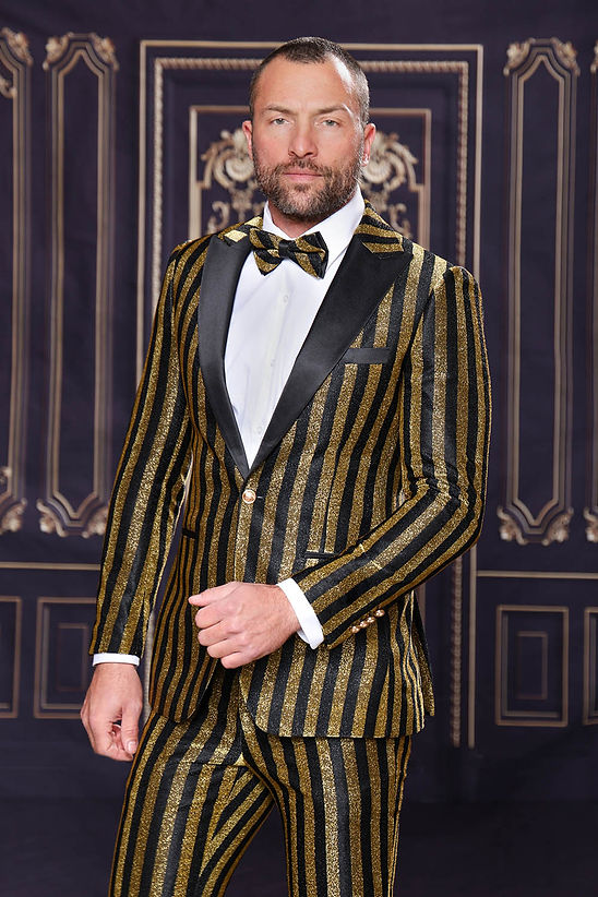A man confidently poses in a Manzini HEATHER Men's Slim-Fit 2-Piece Glitter Stripe Stage Prom Suit in gold, complete with a matching bow tie, standing before a dark decorative background.
