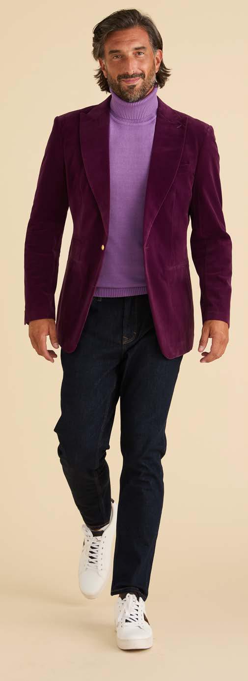 A man wearing an Inserch Velvet Blazer BL007-166 Deep Violet by Inserch, a light purple turtleneck, dark jeans, and white sneakers stands against a plain beige background.