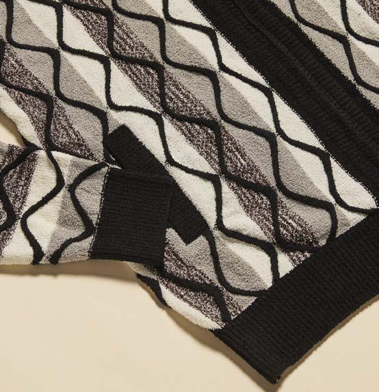 A man stands against a beige background wearing the Inserch Bouclé Yarn Graphic Full Zip Sweater SW614-00001 Black over a light gray turtleneck and black pants.