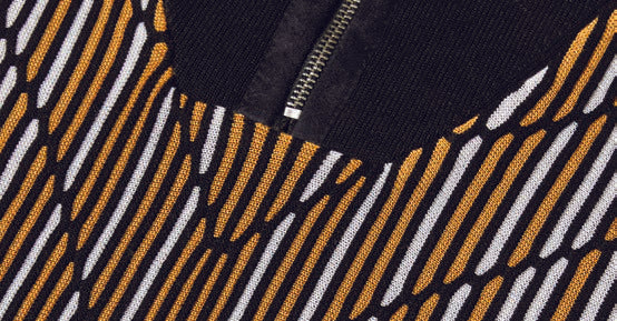 Against a beige background, an individual wears the Inserch Intarsia Panel Half-Zip Sweater SW608-01 Black, which features a black and gold geometric intarsia pattern, paired with yellow pants.