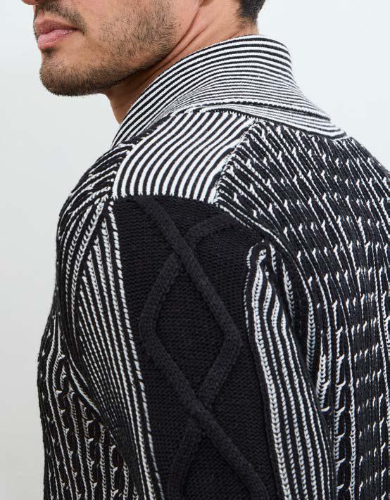A man with short dark hair and facial hair stands against a plain white wall with his hands in his pockets, wearing the Inserch Shawl Collar Cable Sweater SW005-00001 Black by Inserch, crafted from a comfortable cotton-acrylic blend featuring a stylish yarn-dyed pattern.