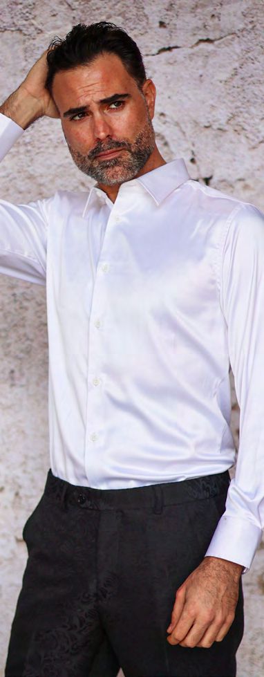 A man with short dark hair and a beard wears the MANZINI INR-101 Slim Fit Satin Spandex White Shirt by Manzini and dark pants, standing in front of a light textured wall with one hand touching his head.