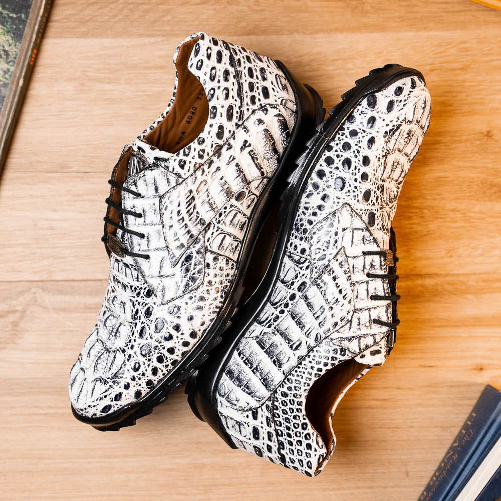 The Marco Di Milano Kratos Newspaper features white and black crocodile-patterned luxury dress shoes with black soles; one shoe stands upright, while the other lies on its side showcasing the hand-painted finish.