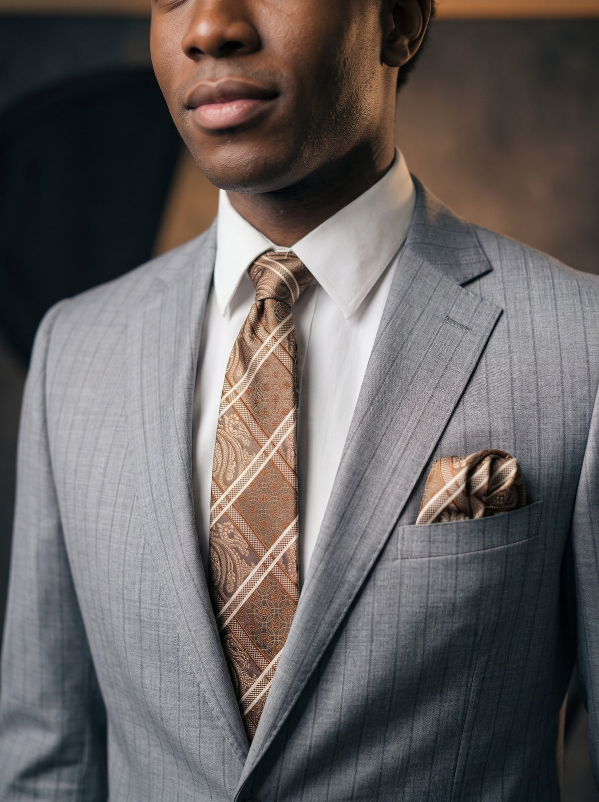 A man wears a Tiglio Luxe Dolcetto Modern Fit Wool Suit TL8093 Grey Pinstripe, styled with a white dress shirt and a tan patterned tie with a matching pocket square.