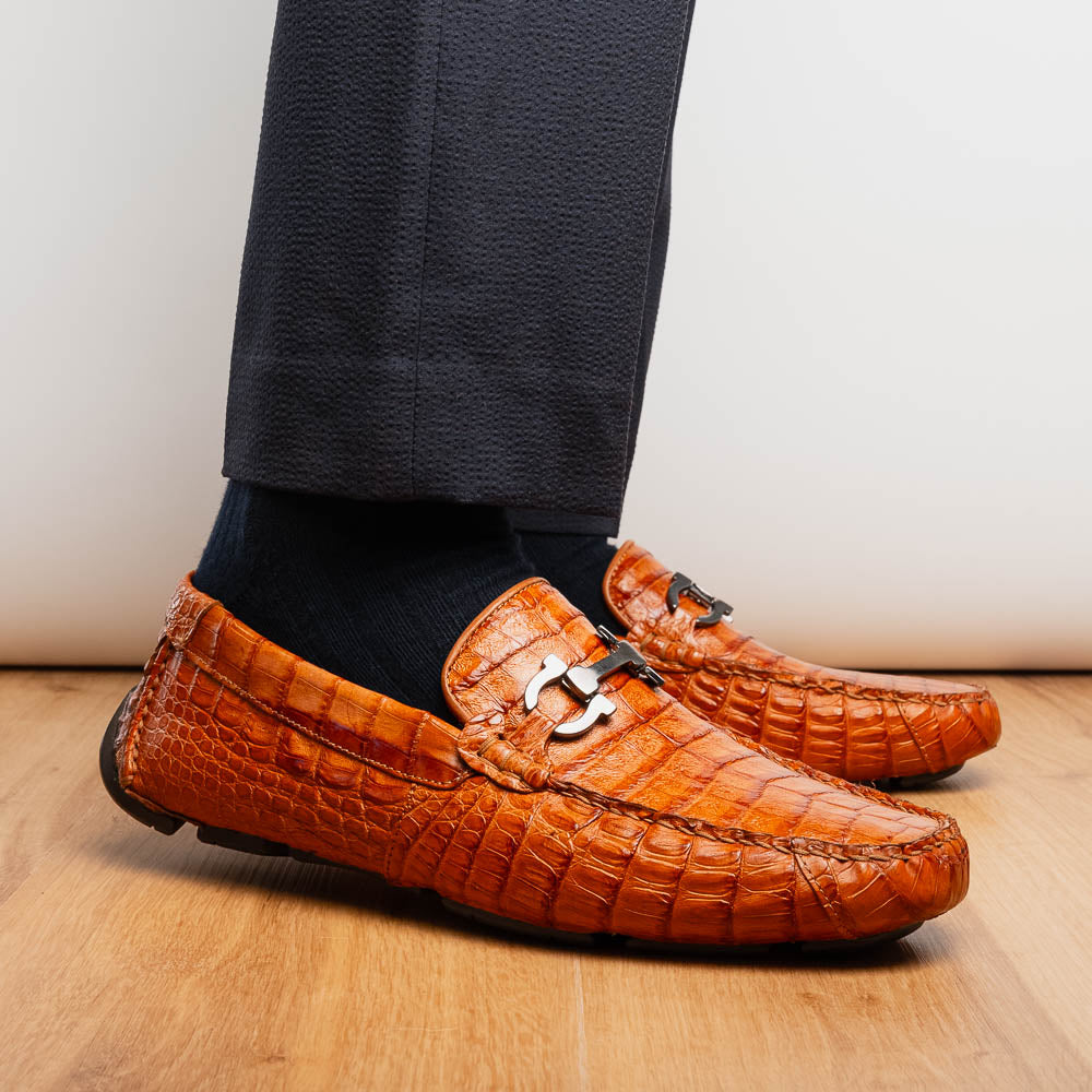 Marco Di Milano’s Parma Brandy loafers feature an orange crocodile pattern, black metal buckles, and hand-painted finishes—one shoe stands upright while the other lies on its side to reveal the detailed sole.