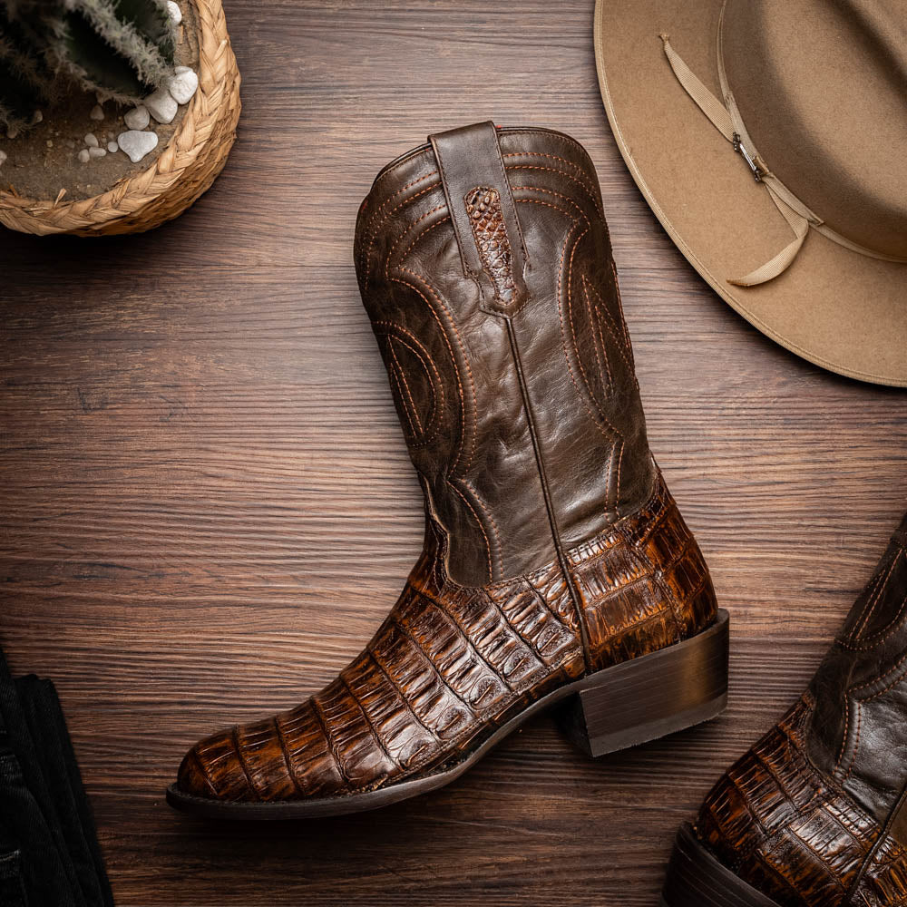 A pair of Marco Di Milano Phoenix Aged Brown men's boots, one upright and one showing the sole, crafted from genuine caiman belly skin with a crocodile texture and detailed stitching—ideal for any Western collection.