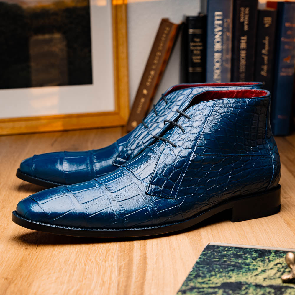 The Marco Di Milano "Pietro Blue Jeans" are luxury men's lace-up boots in blue crocodile-pattern leather, with one standing upright and the other tilted to reveal the tan embossed sole.