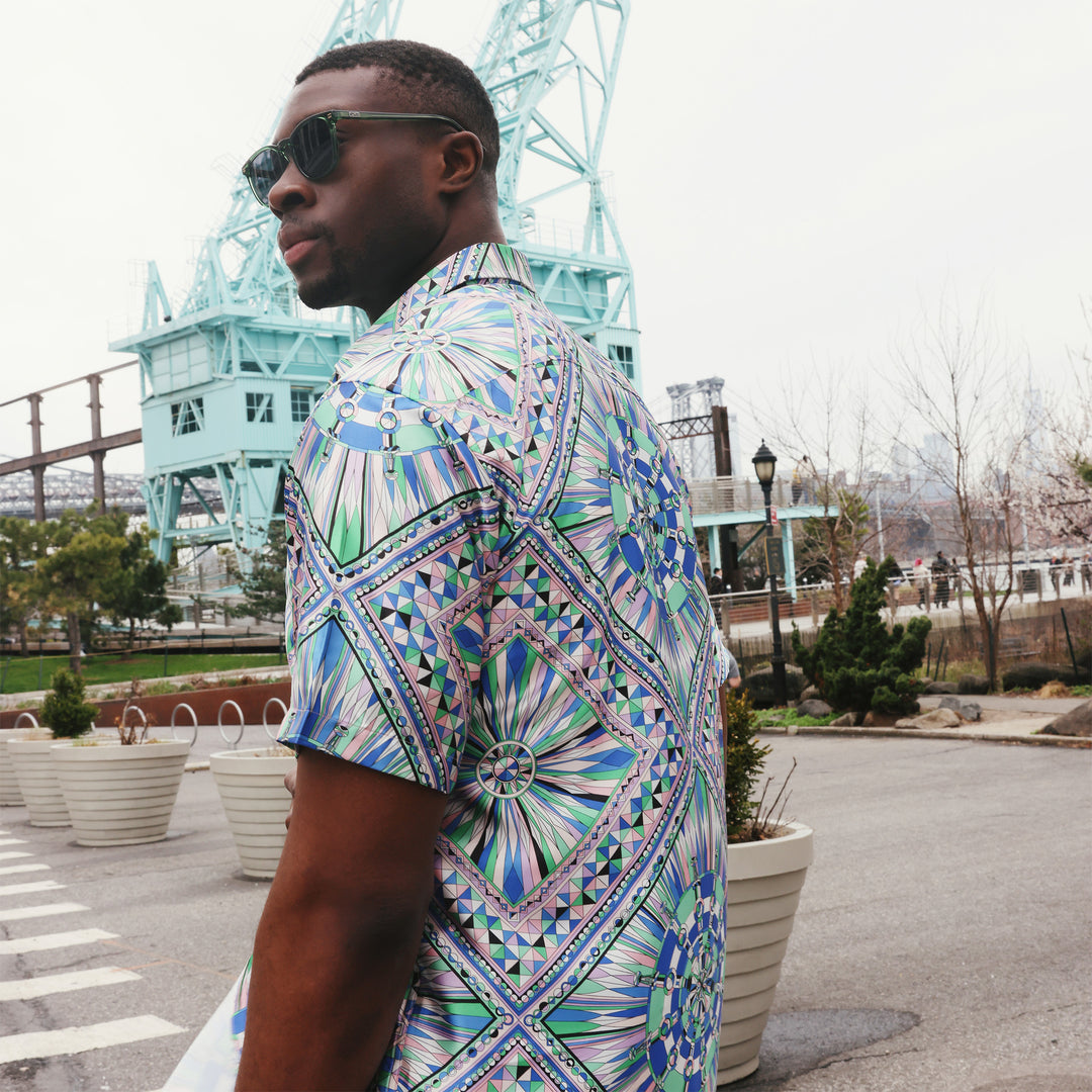 A man wears the Inserch SS Kaleidoscope Print Shirt SS052-00126 Purple by Inserch, featuring a vibrant geometric satin pattern in blue, green, and purple, standing with one hand in his pocket against a beige background.