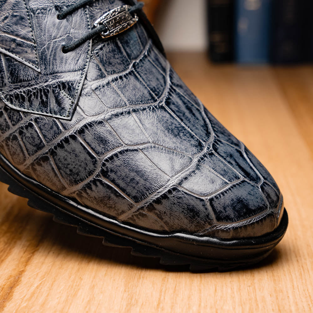 A pair of Marco Di Milano Vulcan Rustic Gray men's dress shoes, showcasing luxury footwear with crocodile-patterned leather, hand-painted finishes, and metal lace accents—one upright, one on its side to display the sole.