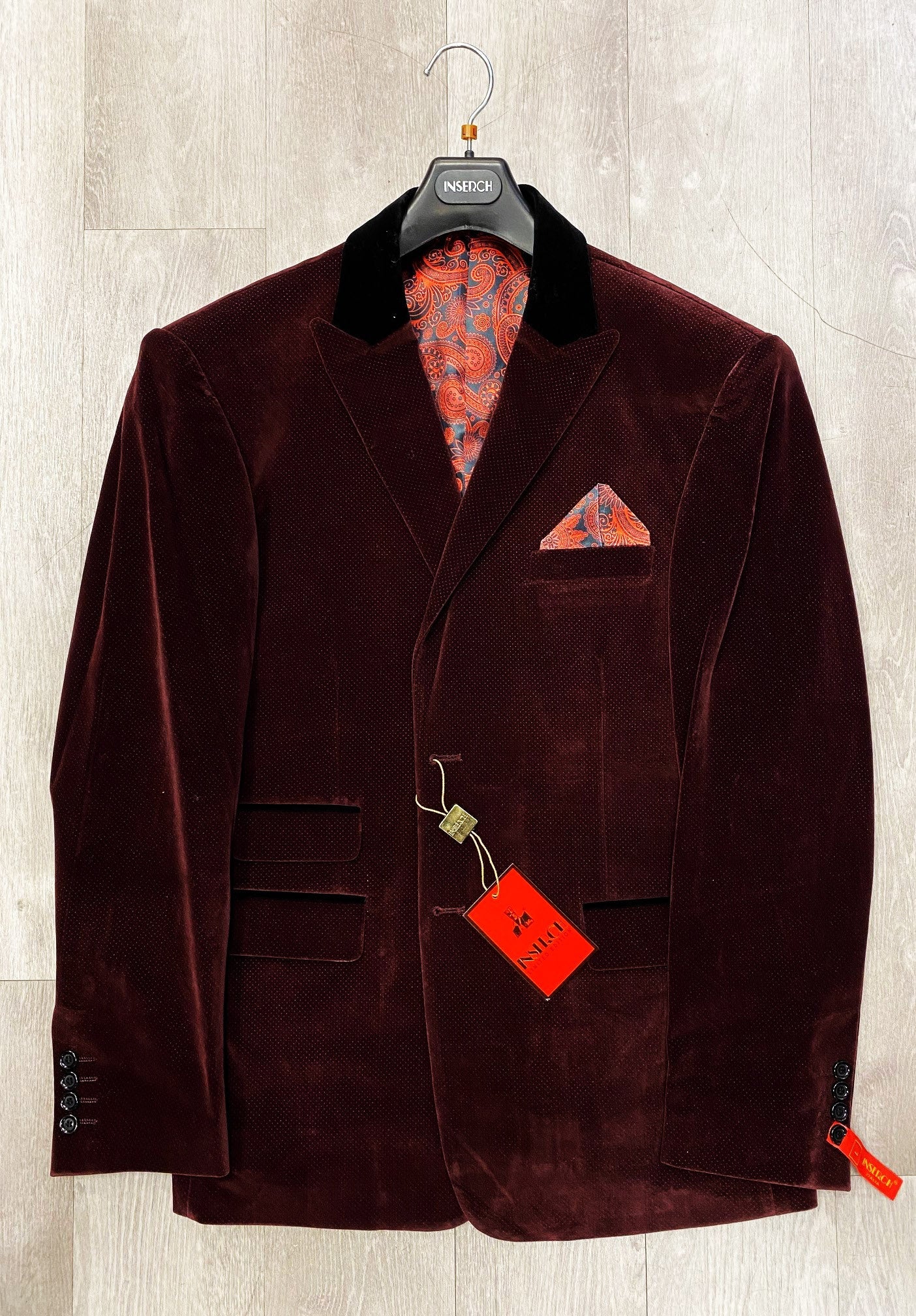 An Inserch Two Button Velvet Dot Print Blazer, in rich dark burgundy with a black velvet collar, paisley lining, and matching pocket square, elegantly displayed on a hanger against a light wood background.
