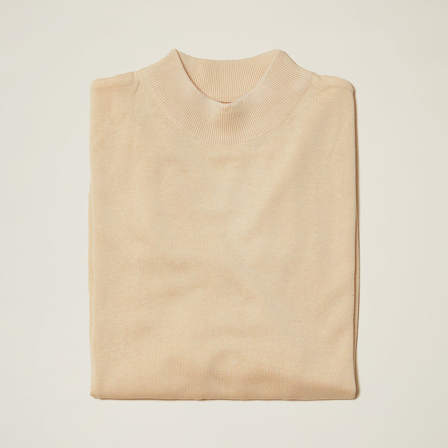 An Inserch Cotton Blend Mock Neck Sweater in Stone (4308-00015) neatly folded, showcasing its ribbed collar, on a plain white surface.