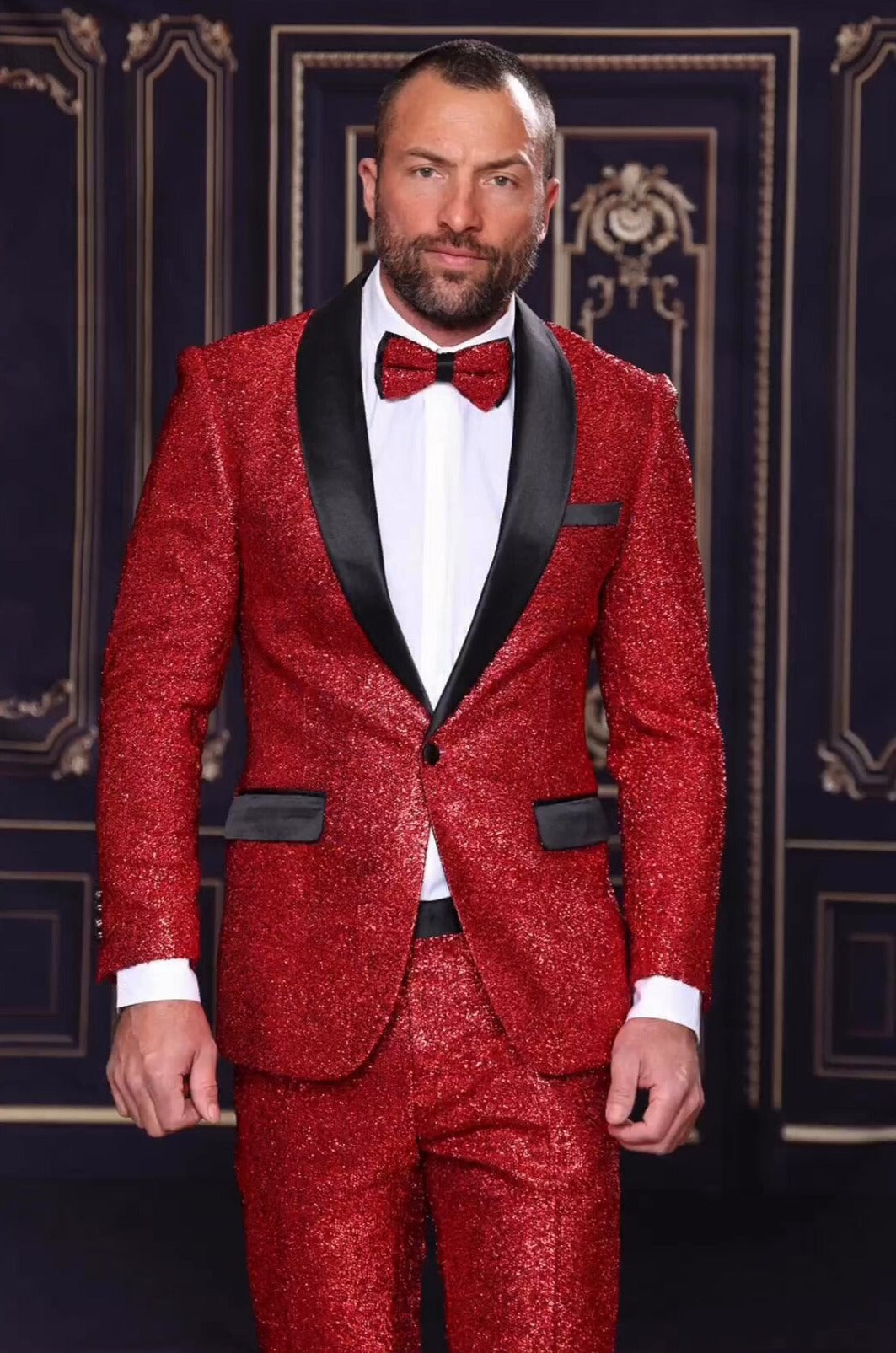 A man dressed in a shimmering Manzini red summer suit, featuring the MANZINI 2pc Slim Fit with a Satin Shawl Collar and paired with a bow tie, stands against an ornate, dark background.