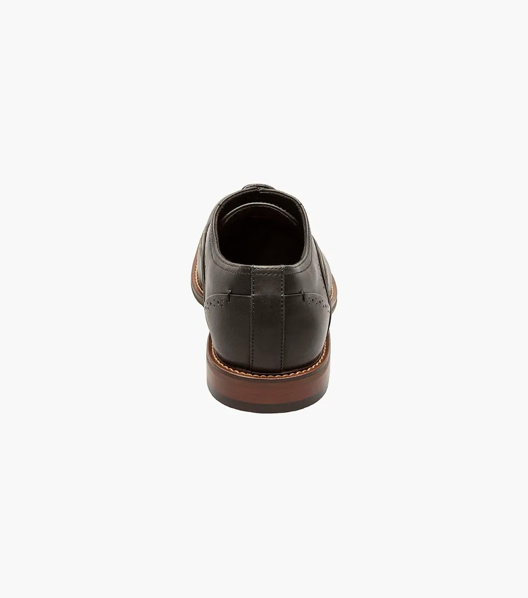 The STACY ADAMS Macarthur Wingtip Oxford (25489-001) is a black burnished leather dress shoe with decorative perforations, brown sole, and black laces, shown against a white background.