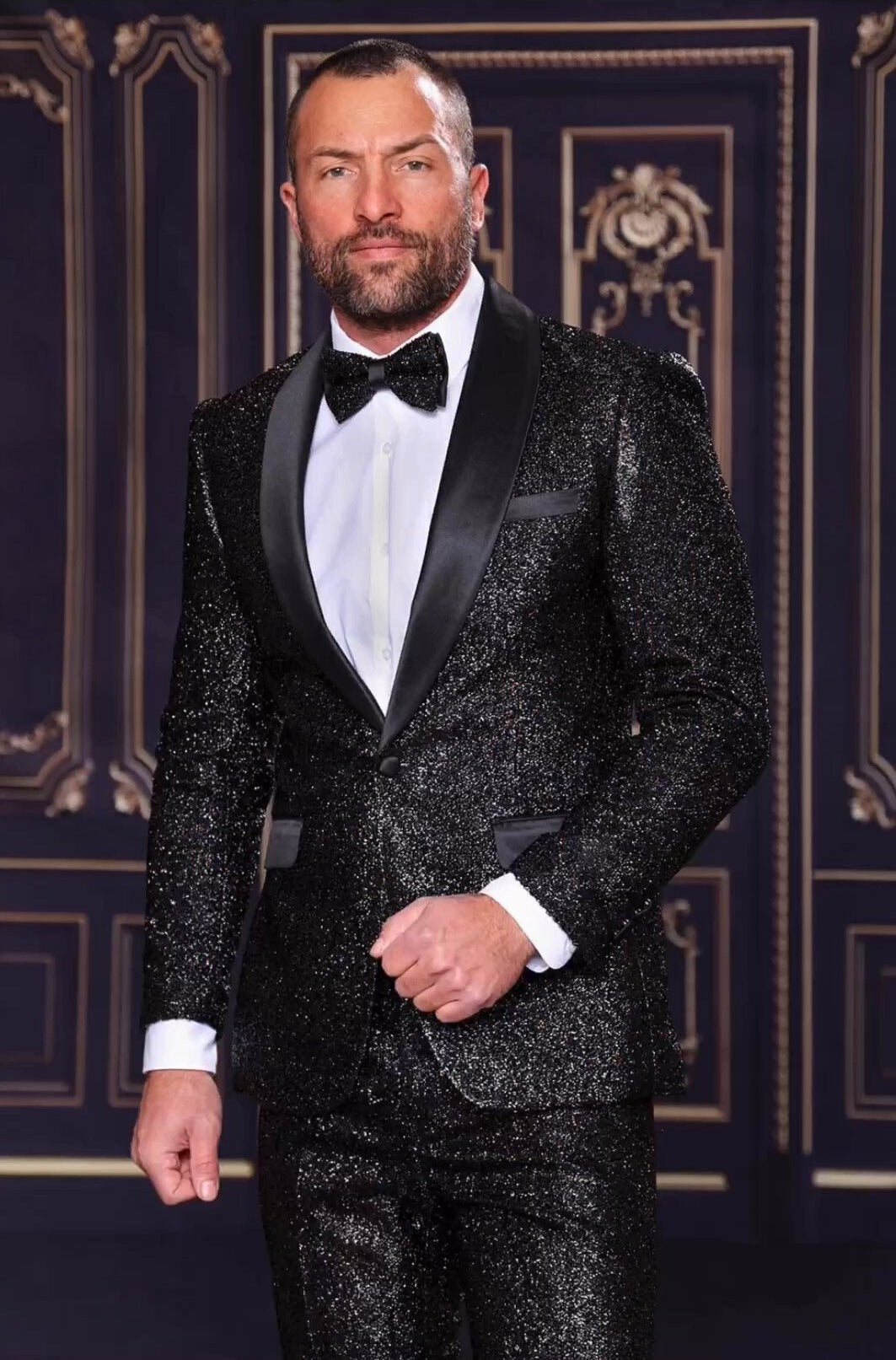 A man wearing the MANZINI 2pc Slim Fit with Satin Shawl Collar Black Suit from Manzini stands against an ornate, dark backdrop, exuding elegance reminiscent of a classic summer suit affair.