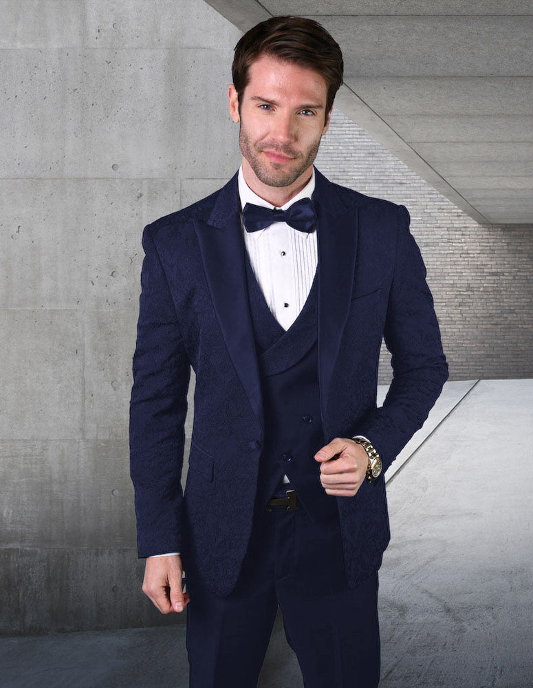 A man wearing the STATEMENT CLOTHING | BELAGIO15-NAVY by Statement Clothing, featuring a tailored fit navy blue tuxedo complete with a bow tie, stands in a concrete hallway with a brick accent wall.