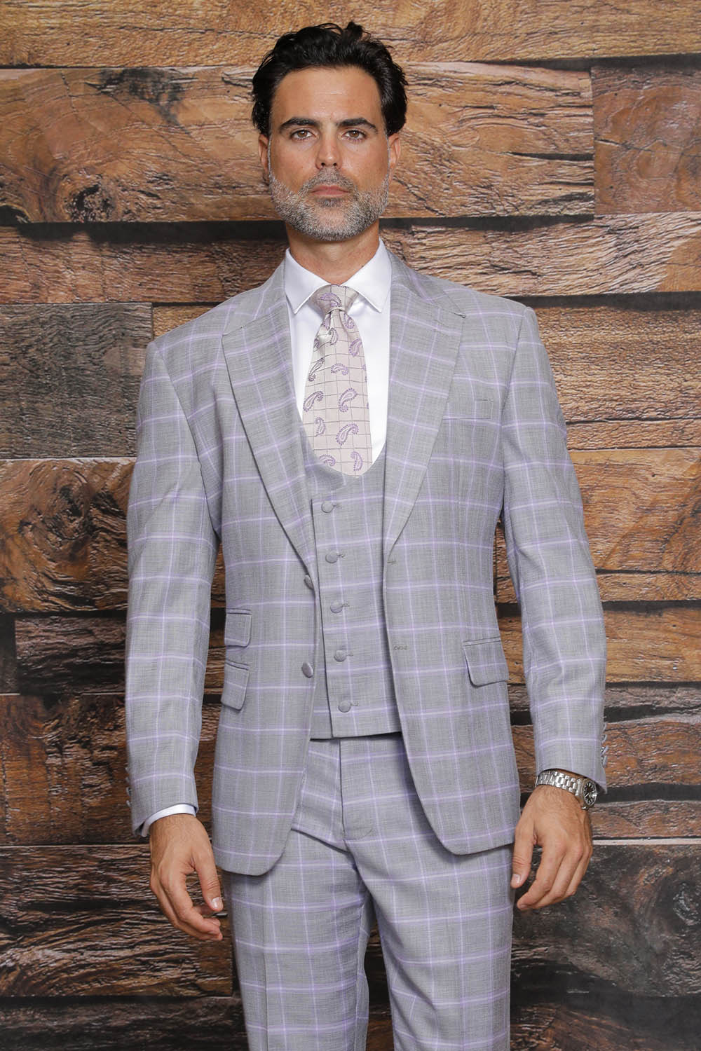 A man dressed in the MANZINI Beverly Ash Suit from Manzini, featuring a gray plaid three-piece design with a paisley tie and modern fit flat front pants, stands against a wooden textured background.