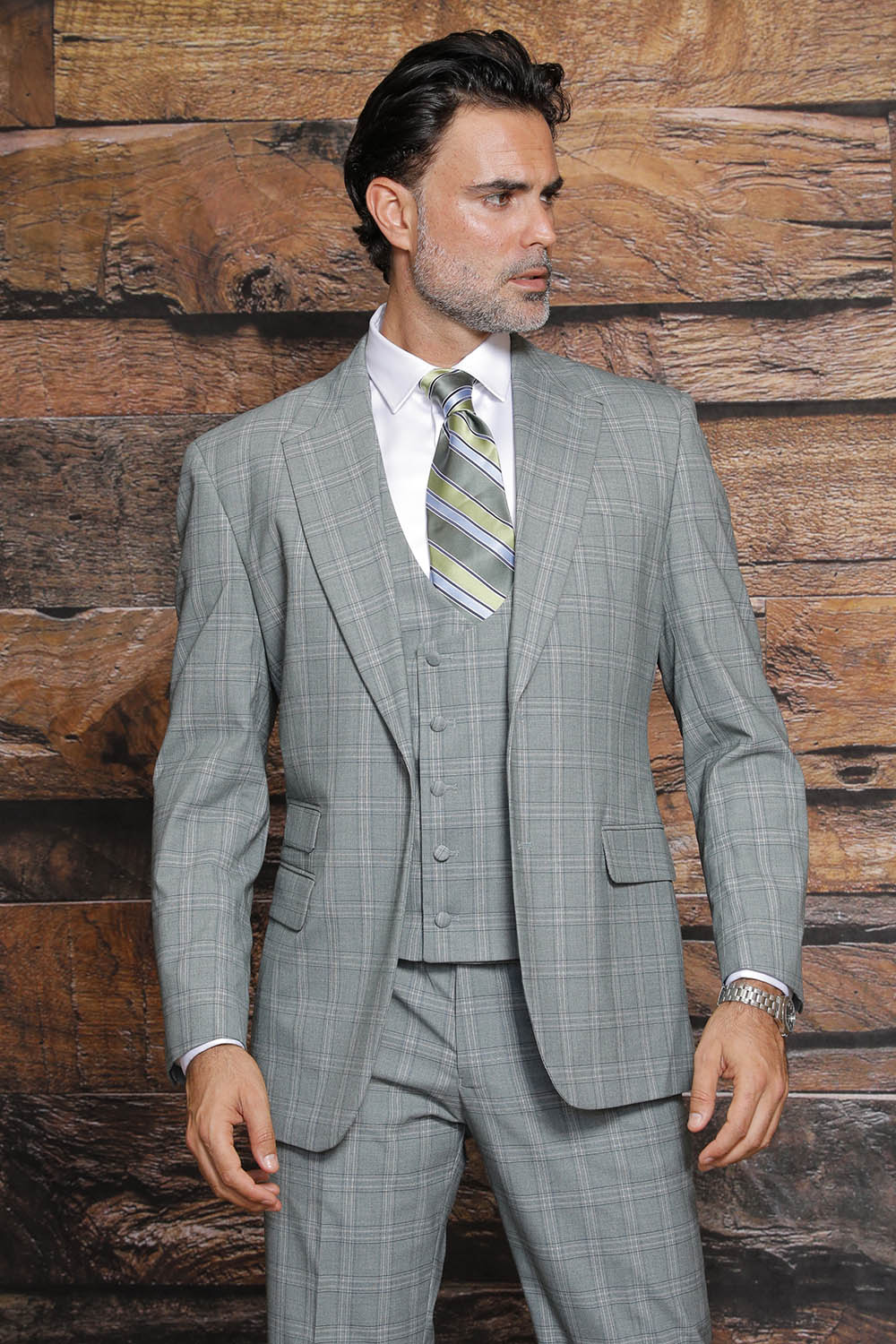 A man wearing the MANZINI Beverly Green Suit by Manzini, featuring a gray plaid design with a modern fit and flat front pants, is complemented by a green striped tie as he stands against a wooden background.