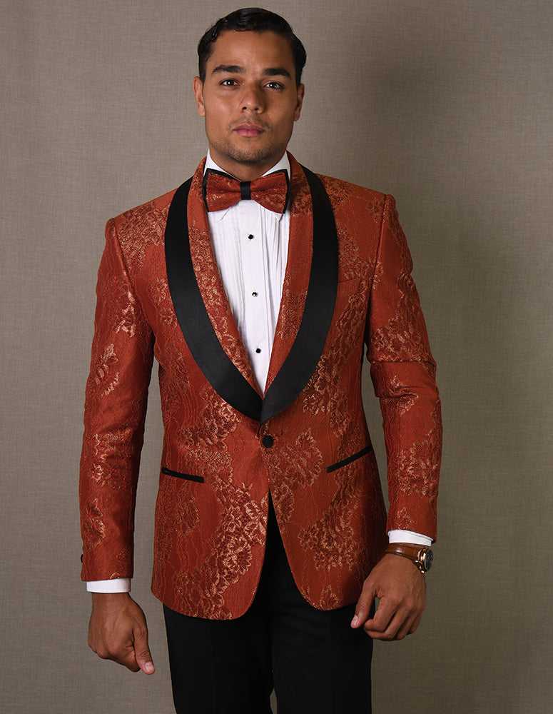 A man wearing the STATEMENT CLOTHING | LJ-104-COPPER tailored fit tuxedo from Statement Clothing, featuring a red patterned design with a black lapel and matching bow tie, stands against a gray background.