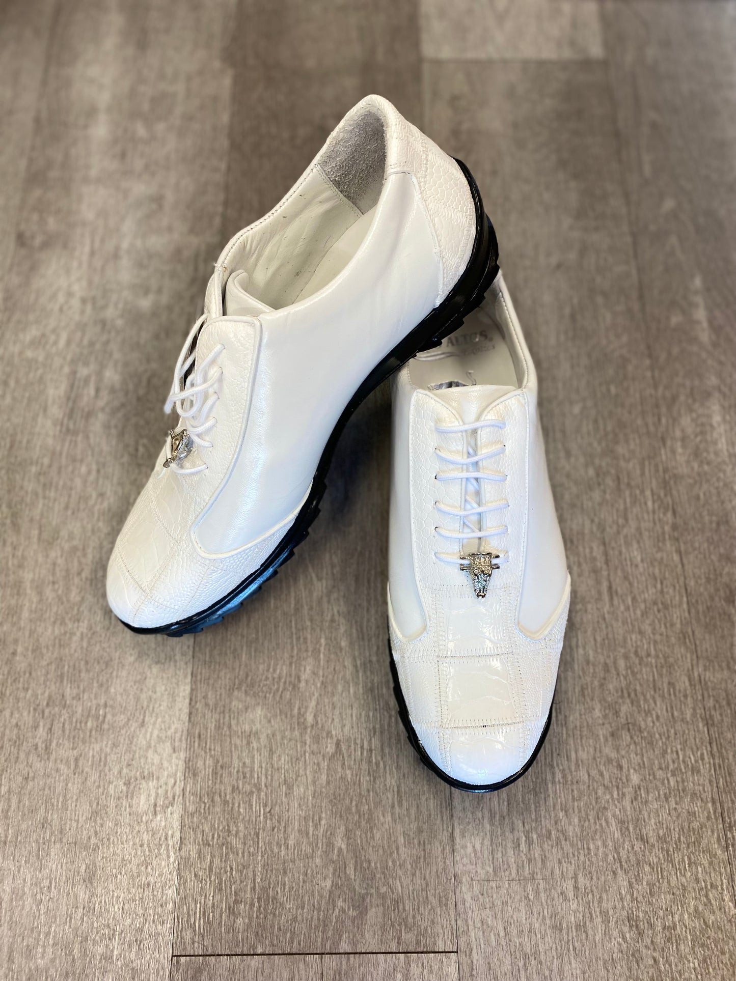A pair of Los Altos ZEUS White Ostrich Leg shoes with black soles embellish the gray wooden floor. Crafted by Los Altos, these shoes are made from elegant ostrich leg leather, featuring a subtle pattern and laces that epitomize refined leather footwear.