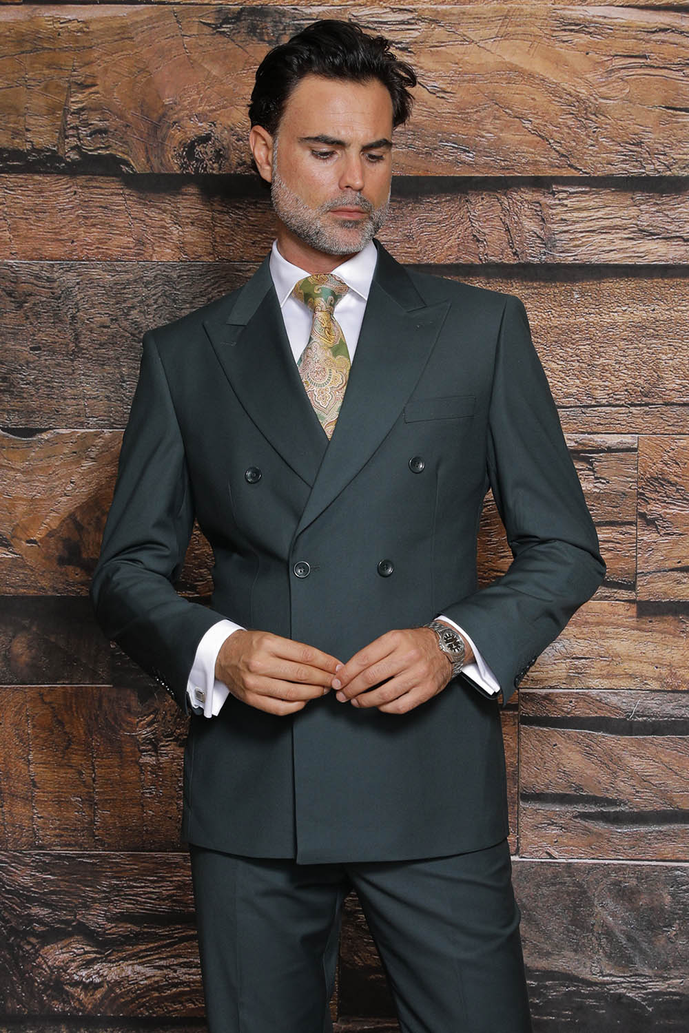 A man wearing a Manzini MANZINI Luna Hunter Suit, featuring a dark green double-breasted design and a modern fit, along with a patterned tie, stands against a wooden background.