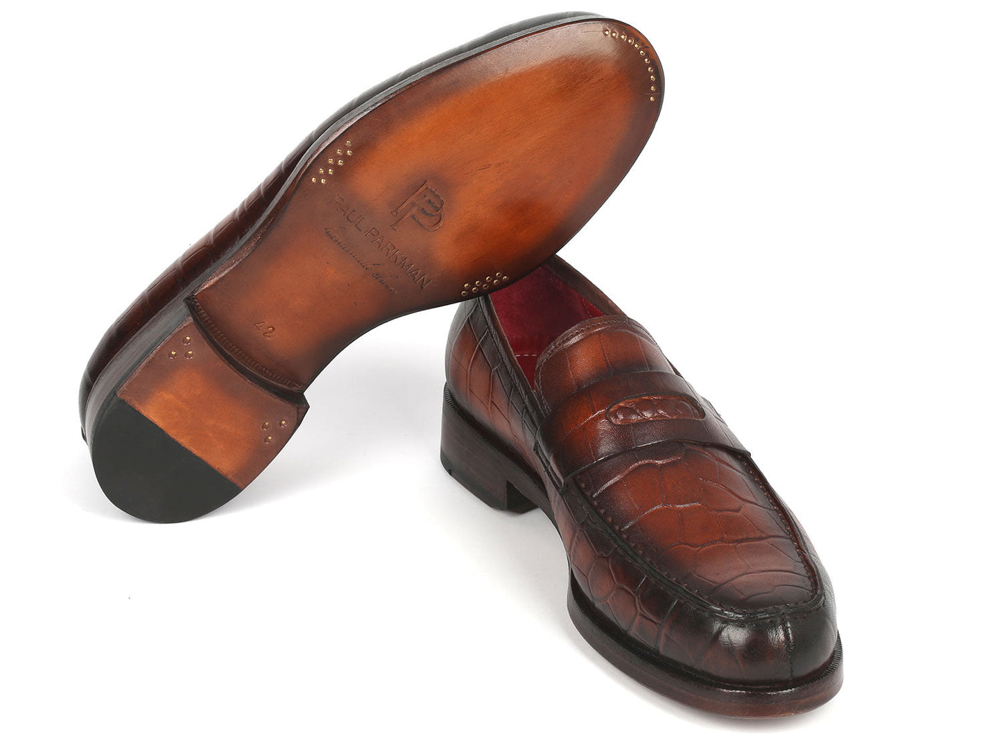 The Paul Parkman Brown Crocodile Embossed Calfskin Penny Loafers - 3857-BRW, featuring a stylish red interior lining, are elegantly displayed with one shoe resting at an angle on the other.