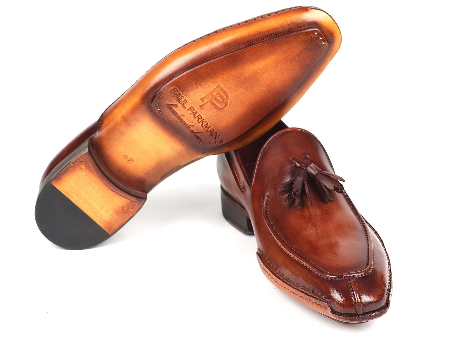 The Paul Parkman Hand-Sewn Tassel Loafers Brown - 082-BRW, expertly crafted from hand-painted leather, exude a sleek, glossy finish. One shoe stands upright as the other leans gracefully against it, accentuating their sophisticated appeal.
