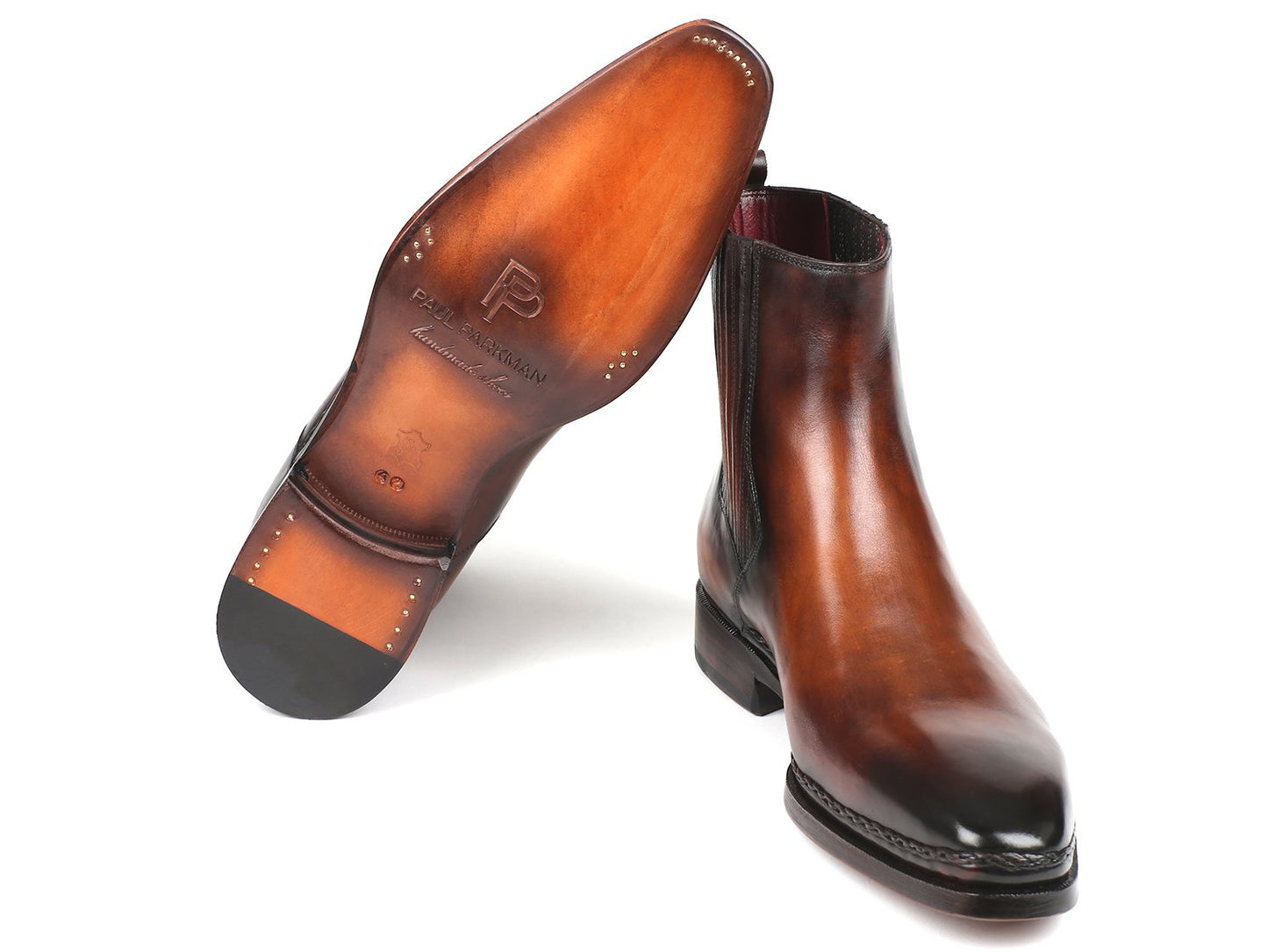 The Paul Parkman Chelsea Boots in Brown Burnished Leather (BT57-BRW) are expertly handmade with calfskin and feature elastic side panels along with low heels. One boot stands upright while the other is tilted slightly, showcasing their polished design.