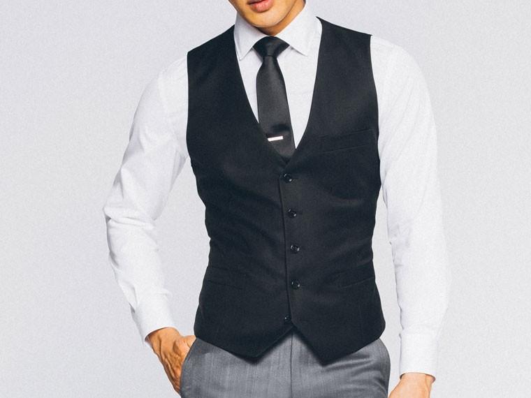A person wearing the Vinci Suits Slim Fit Single Breasted 5 Button Vest (Black) OV-900 over a white shirt with a black tie and gray pants stands against a plain background.
