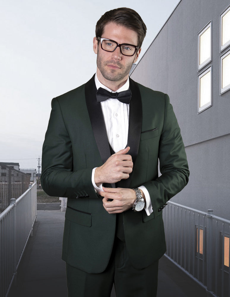 A man in a dark suit with a bow tie and glasses stands on a walkway, adjusting his sleeve. His STATEMENT CLOTHING | TUX-SH-HUNTER tuxedo from Statement Clothing exudes elegance, accentuated by the classic style of flat front pants.