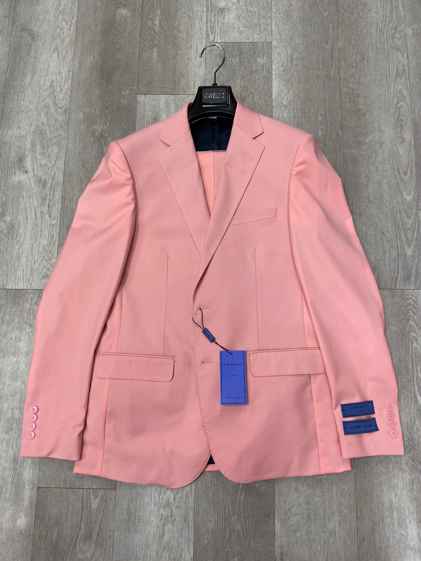 The Cavelli Uomo Porto SLIM FIT Suit 3488/2 in pink by CAVELLI features a notched lapel, two front pockets, and lightweight slim fit. Displayed on a hanger over a wood floor with tags attached to the sleeve and buttonhole. FINAL SALE—NO RETURNS OR EXCHANGES.