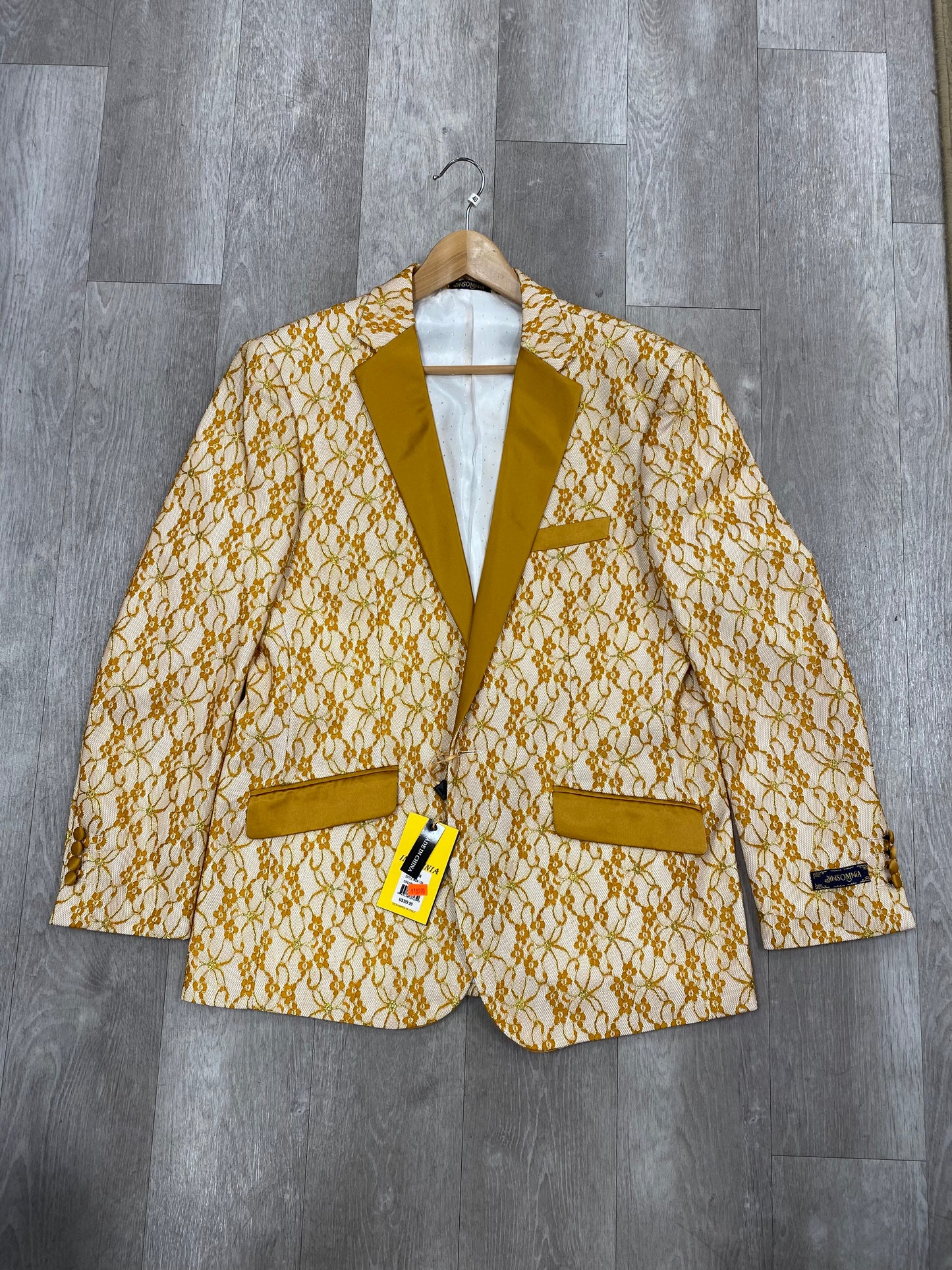 The Insomnia Yellow Fashion Blazer by Inserch, featuring a gold and white pattern with mustard-colored trim and pockets, is elegantly displayed on a wood floor. Please note that it is an item of final sale with no returns or exchanges available. Available in sizes L - M.
