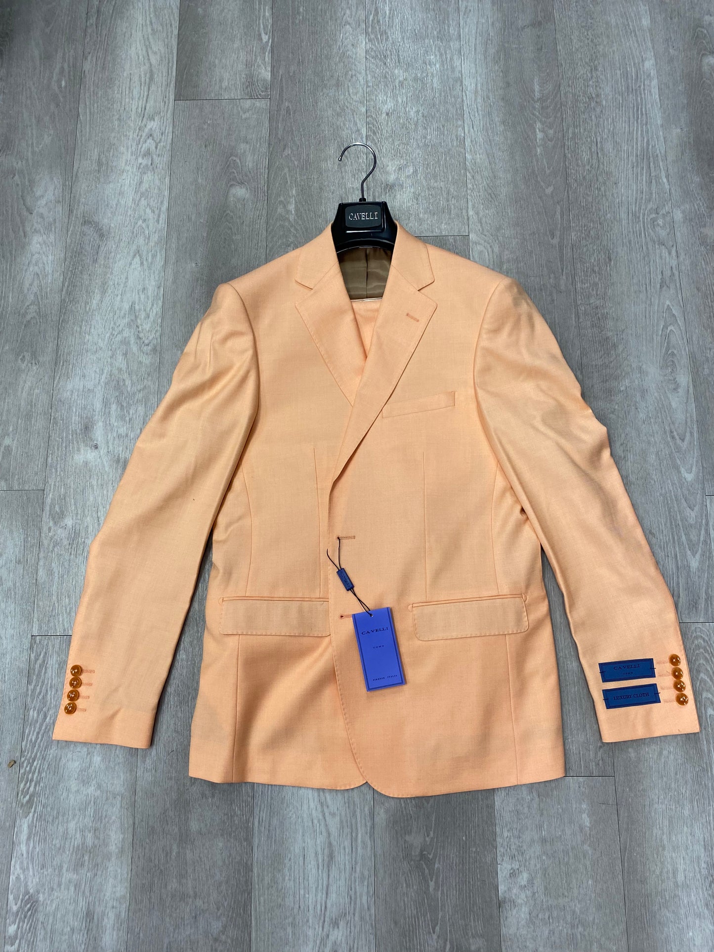 The Cavelli Uomo Porto SLIM FIT Suit 1986/11 in light orange by CAVELLI is shown on a hanger against a gray wood floor, featuring gold buttons and a blue tag. FINAL SALE: no returns or exchanges. Ideal for office wear.