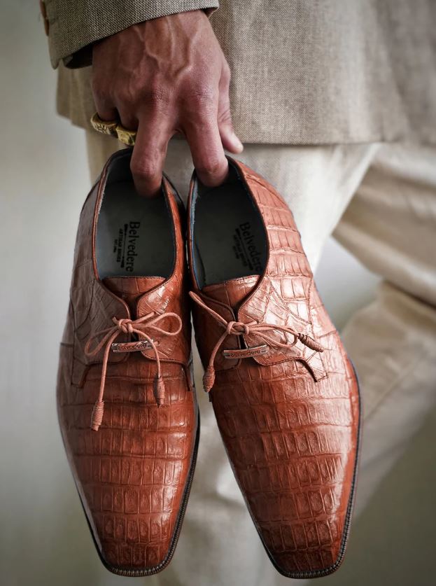 A person in a beige suit holds a pair of brown crocodile leather dress shoes by the laces.