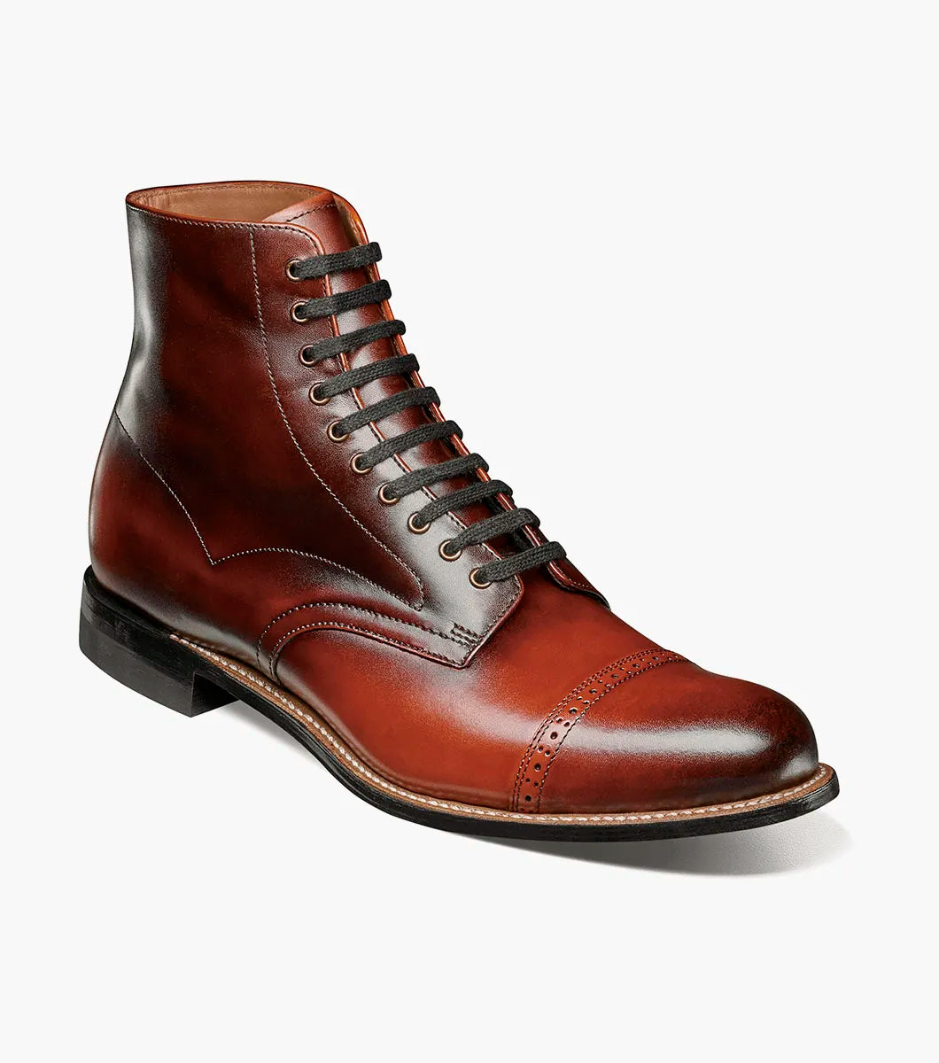 The Stacy Adams Madison Cap Toe Lace Up Boot in Cognac (00110-221) features brown leather, black laces, brogue detailing, and a stacked heel. This stylish STACY ADAMS boot is shown on a white background.