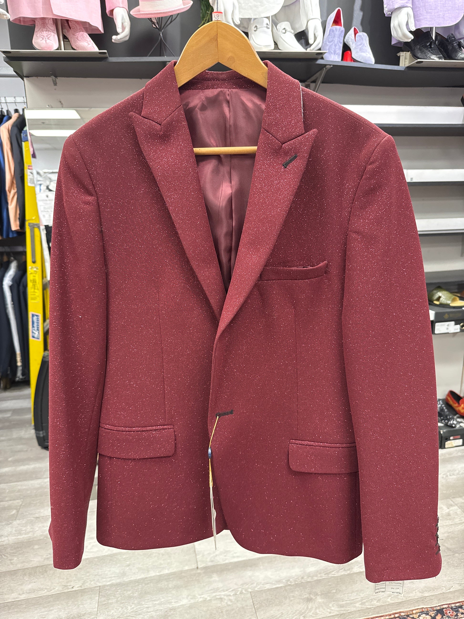 The EJ SAMUEL JACKET 19 CLEARANCE, featuring a maroon hue and notched lapel, is displayed on a wooden hanger in a clothing store with shelves and other garments visible in the background; please note this item is final sale and cannot be returned or exchanged.