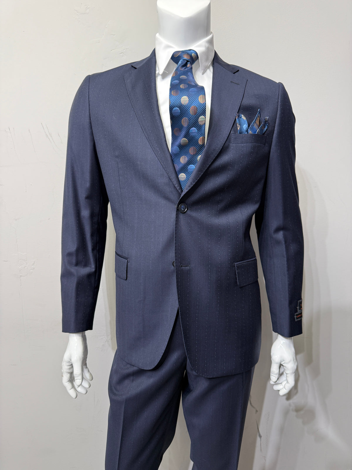 A mannequin dressed in the Tiglio Luxe Dolcetto Modern Fit Navy Fancy Pinstripe Wool Suit TL8107 by Tiglio Luxe, paired with a crisp white dress shirt, a blue polka dot tie, and a coordinating pocket square, posed against a plain light-colored background, exemplifies the refined elegance of tailored men’s clothing.