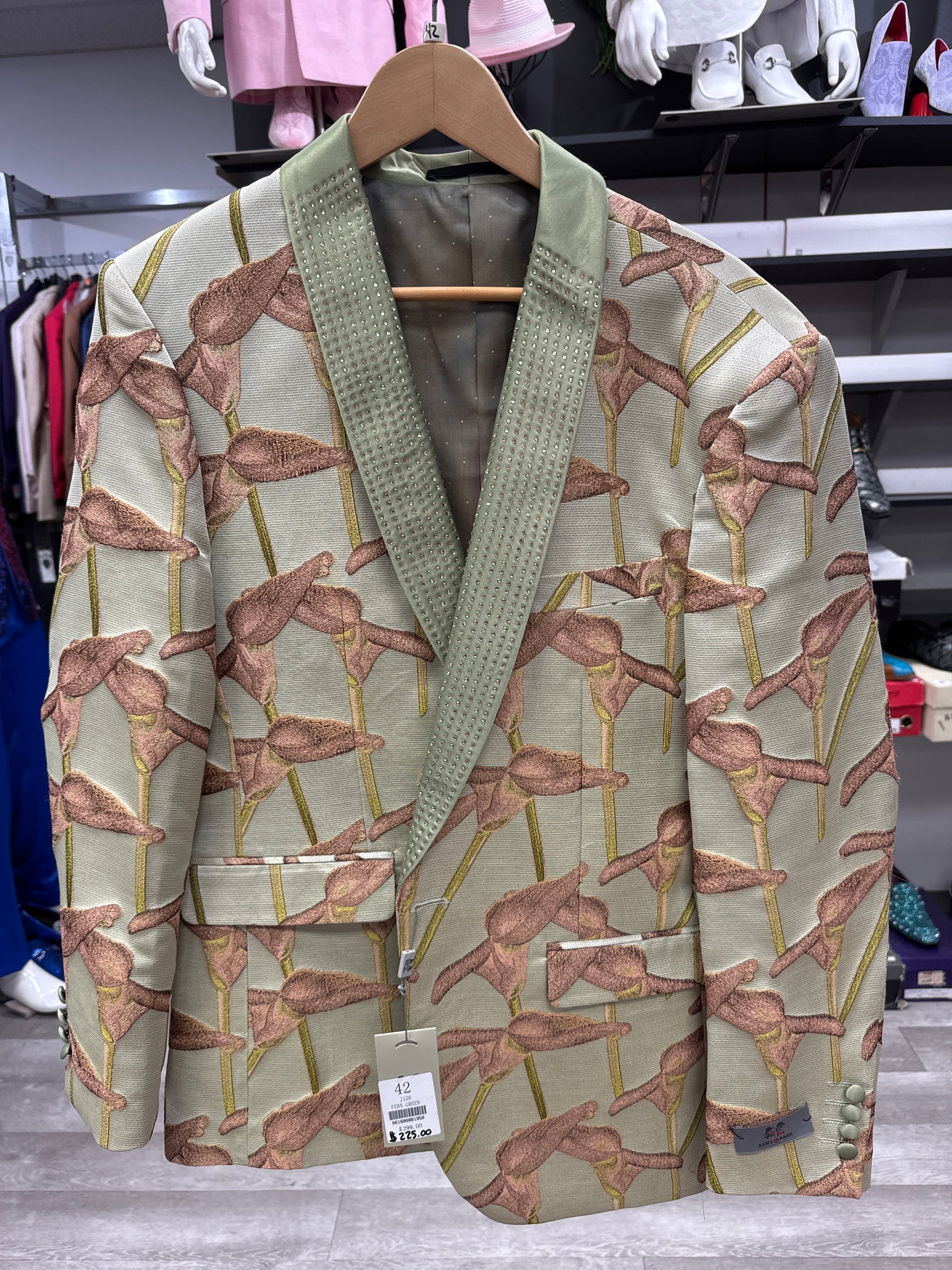 An EJ SAMUEL JACKET 22 CLEARANCE—a light green blazer featuring a flamingo pattern and a textured, shiny green shawl collar—hangs on a rack in the clothing store. Please note: this is a final sale item and cannot be returned or exchanged.