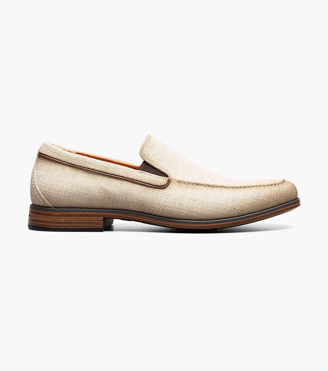 The Stacy Adams Ridgely Moc Toe Slip On (Natural) features a textured fabric upper, brown trim, anatomical arch support, and a low wooden heel for comfort, shown here on a white background.