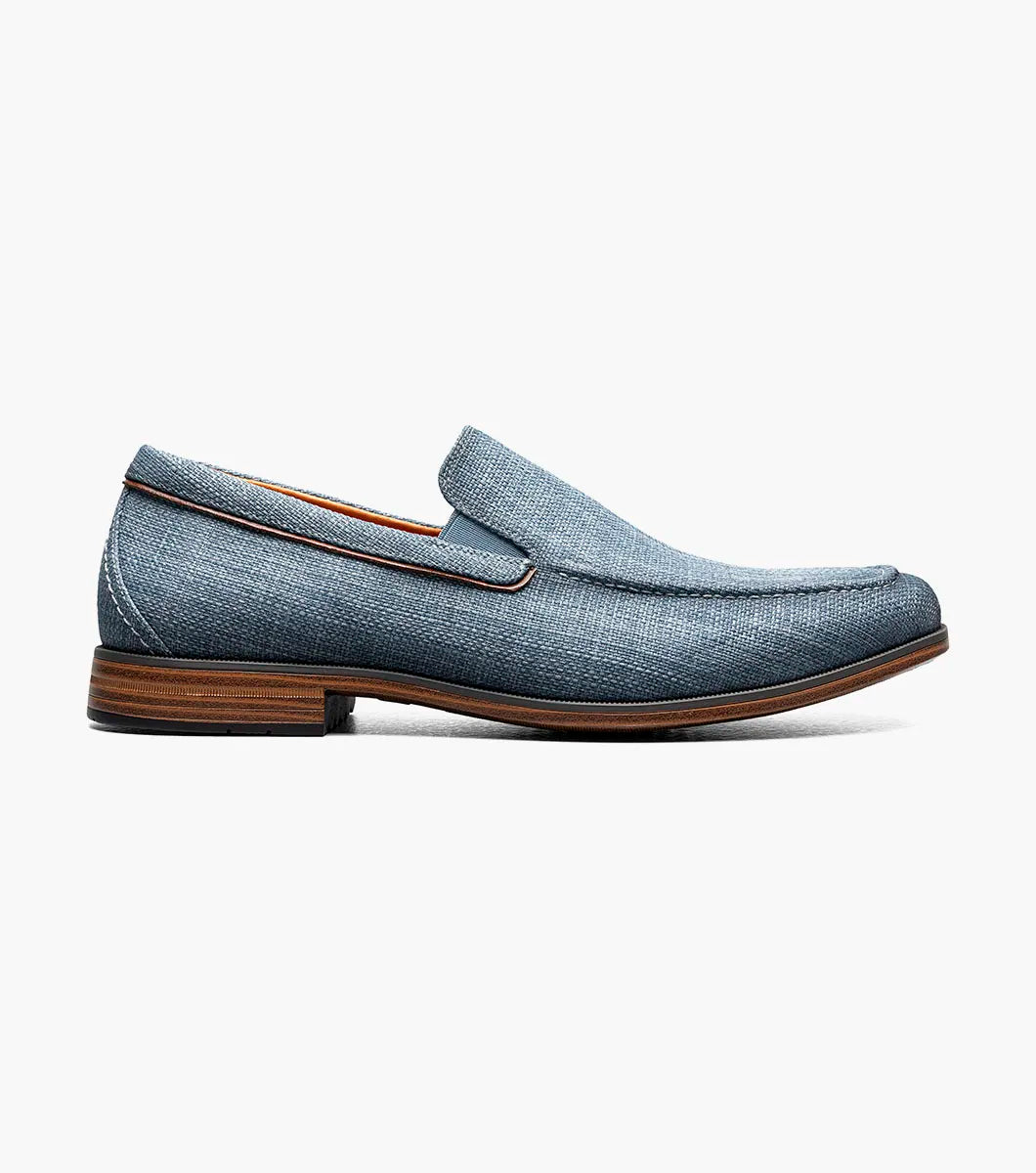 The Stacy Adams Ridgely Moc Toe Slip On in navy features a textured fabric upper, breathable leather lining, brown trim, and a low stacked wooden heel, shown against a white background.