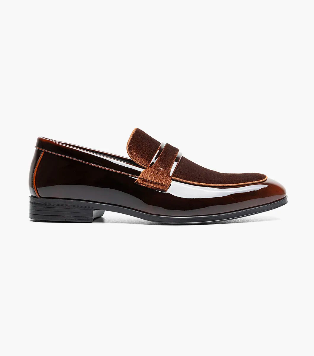 The Stacy Adams Spratley Moc Toe Saddle Slip On (Brown, 25686-200) is a men’s loafer with a glossy leather finish, cushioned insole, and velvet strap detail, displayed angled on a plain white background.