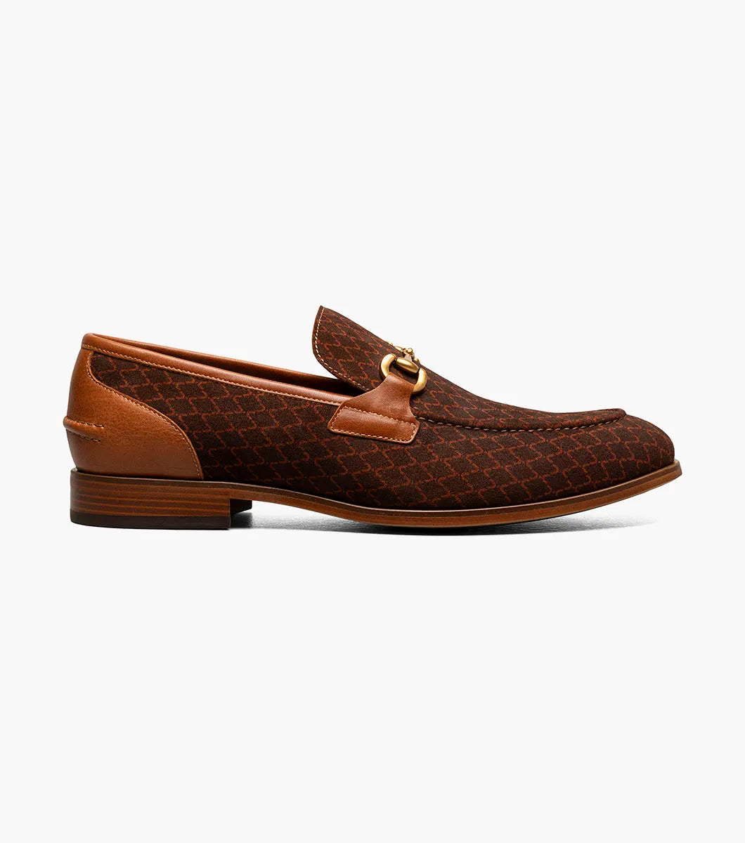The Stacy Adams Brinkley Moc Toe Bit Slip On in tan features a patterned fabric upper, leather trim, gold metal bit ornament, and a low wooden heel, displayed on a white background.