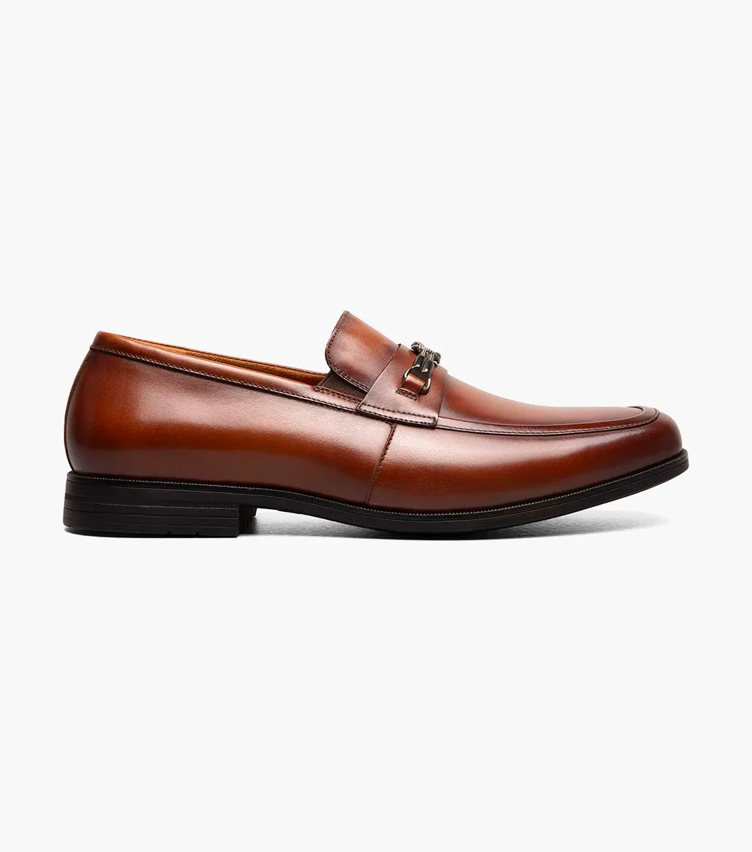 The Stacy Adams Rellaire Moc Toe Bit Slip On in cognac features a brown leather upper, low heel, decorative metal bit, cushioned footbed, and black sole. Displayed on a plain white background.