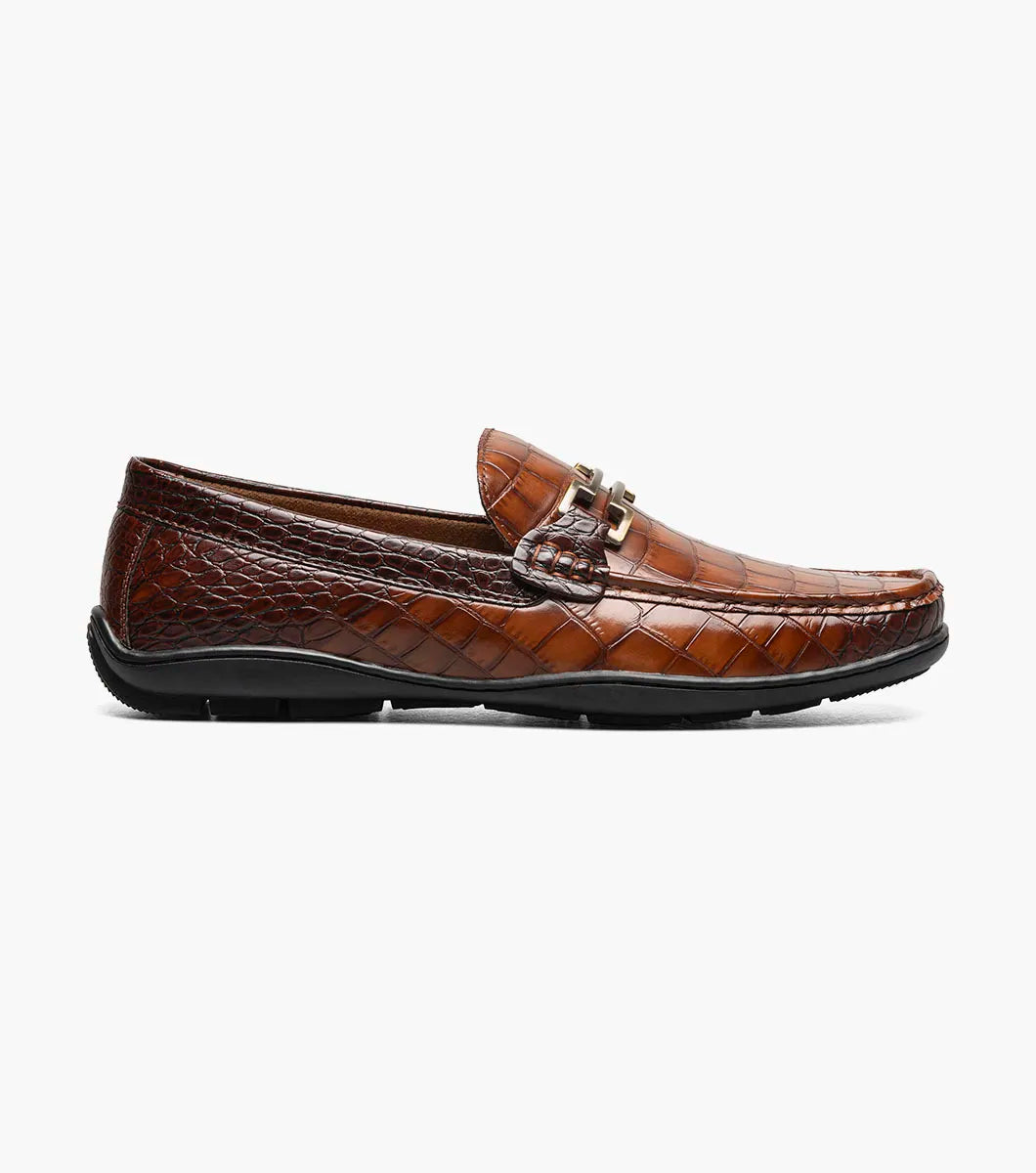The STACY ADAMS Embrey Moc Toe Bit Slip On in Cognac (25703-221) features brown crocodile print leather, a metallic buckle, a cushioned footbed for comfort, slip-on design, and is showcased against a plain white background.