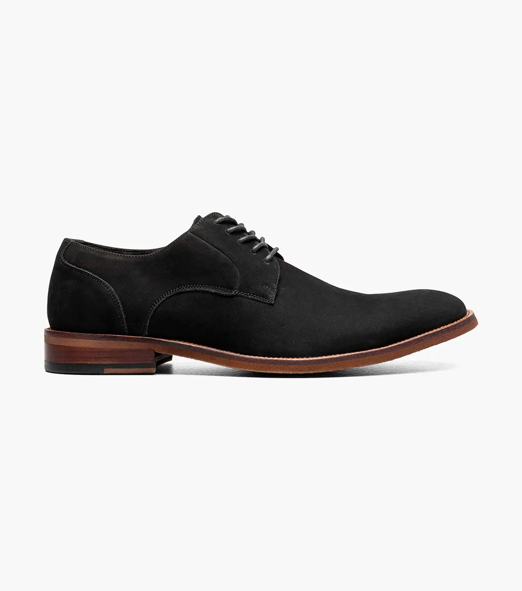 The Stacy Adams Preston Plain Toe Lace Up (25650-001) is a men’s tumbled leather dress shoe in black, featuring black laces, a brown wooden sole, and simple stitching. Displayed elegantly on a white background.