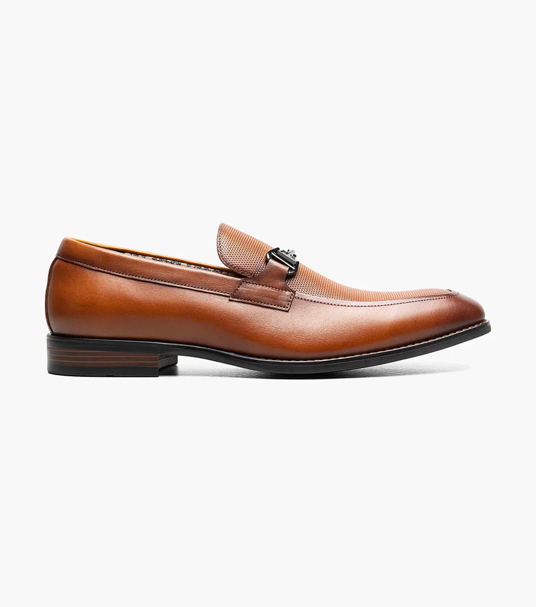 The Stacy Adams Lawton Moc Toe Bit Slip On in Cognac (style 25796-221) features a textured brown leather upper, classic moc toe slip-on design, cushioned footbed, silver metal bit detail across the vamp, and a low stacked heel. Shown at an angle on a white background, this STACY ADAMS shoe combines comfort with timeless style.