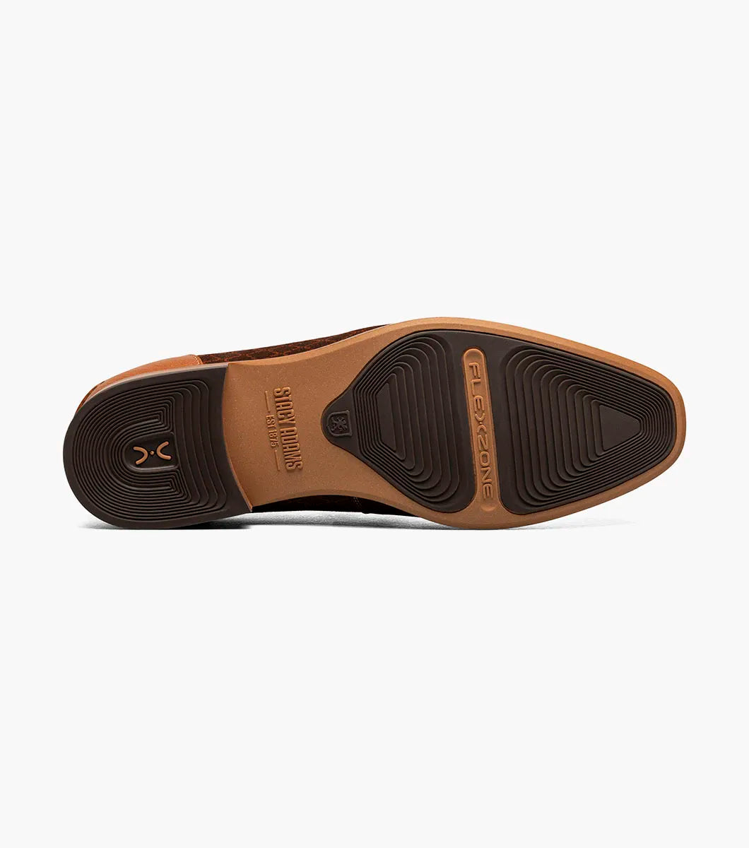 The Stacy Adams Brinkley Moc Toe Bit Slip On in tan features a patterned fabric upper, leather trim, gold metal bit ornament, and a low wooden heel, displayed on a white background.